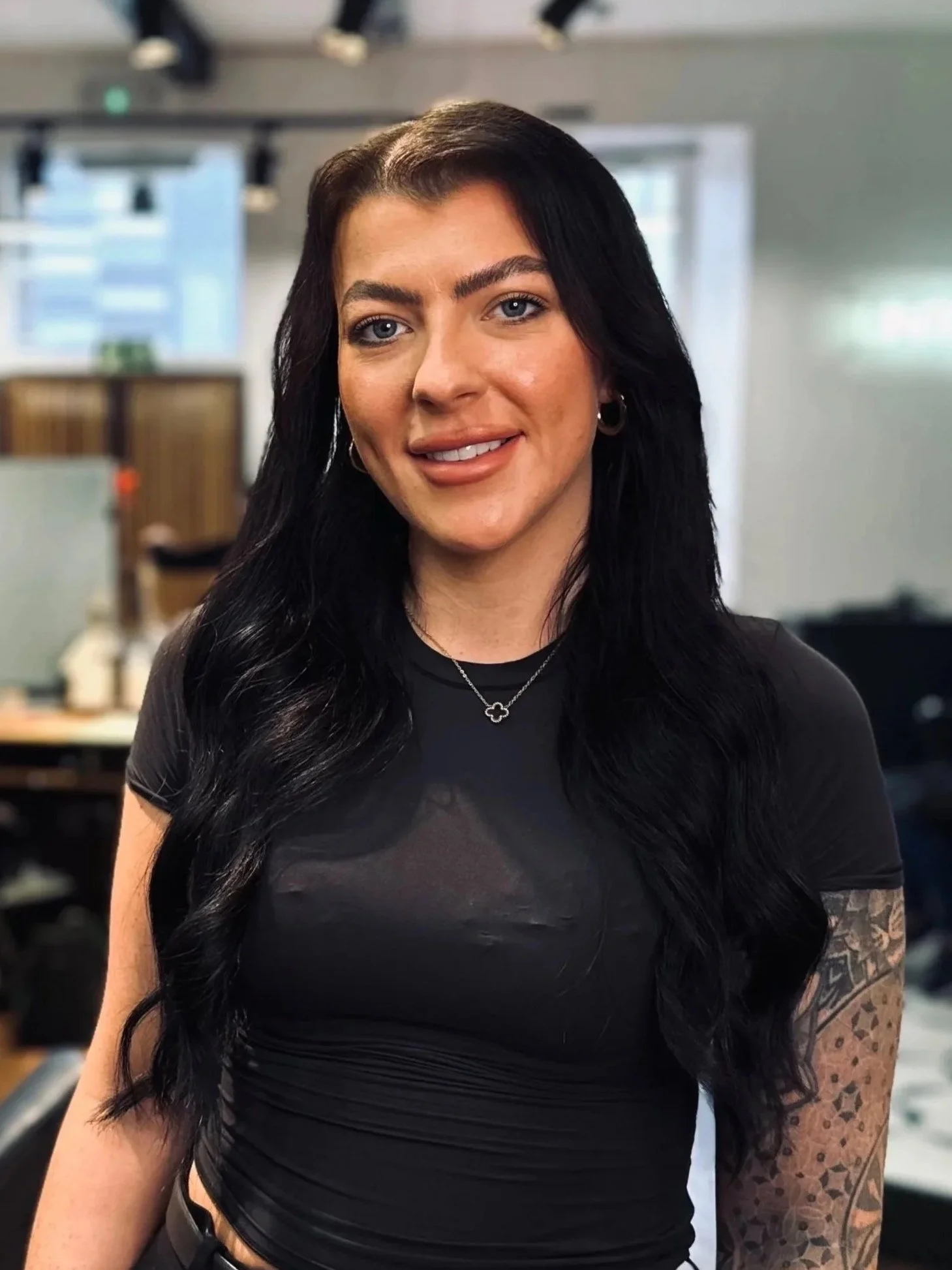 A woman with long black hair, wearing a black top and black pants with a belt, standing in an office environment with people working in the background.