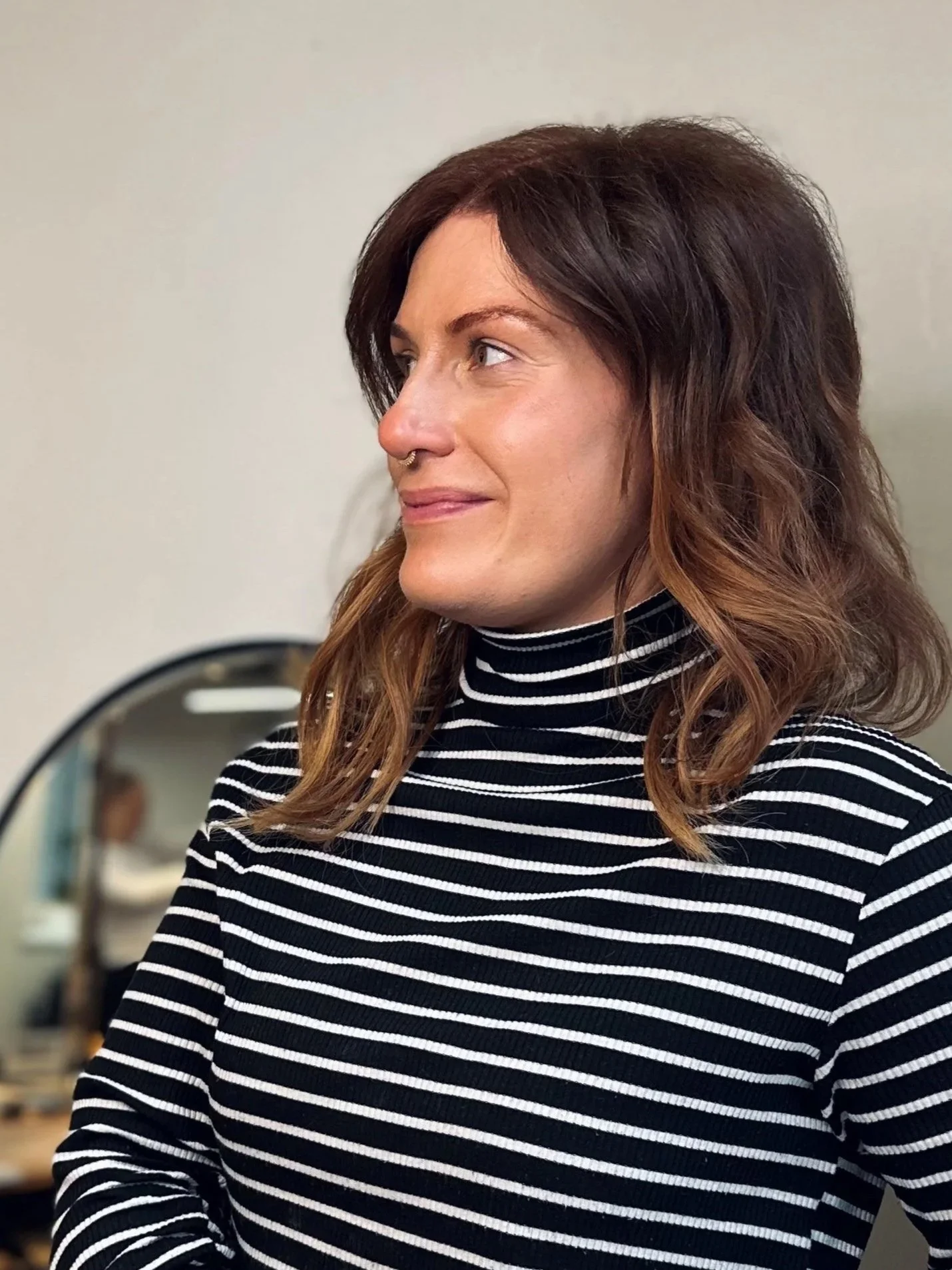 A woman with shoulder-length brown hair and tattoos on her arms, wearing a black and white striped long-sleeve shirt, standing indoors near a mirror.