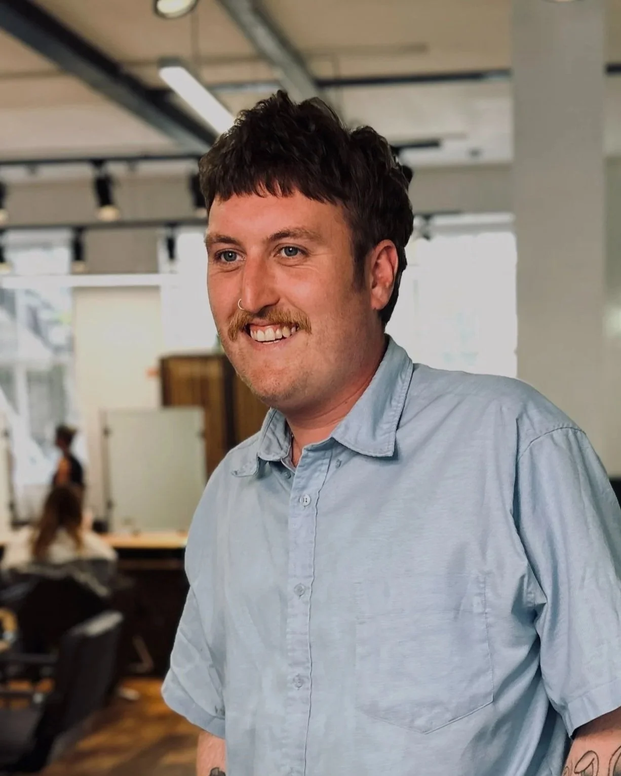 A man with a mustache and tattoos, wearing a light blue shirt, standing in a modern office or coworking space with wooden floors, working station desks, and other people in the background.