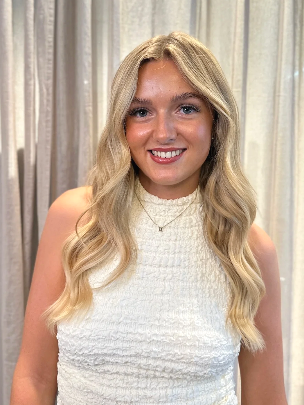 A young woman with blonde hair, wearing a black sleeveless top and white textured pants, standing and smiling in an indoor office or coworking space with wooden floors, tables, chairs, and large windows.