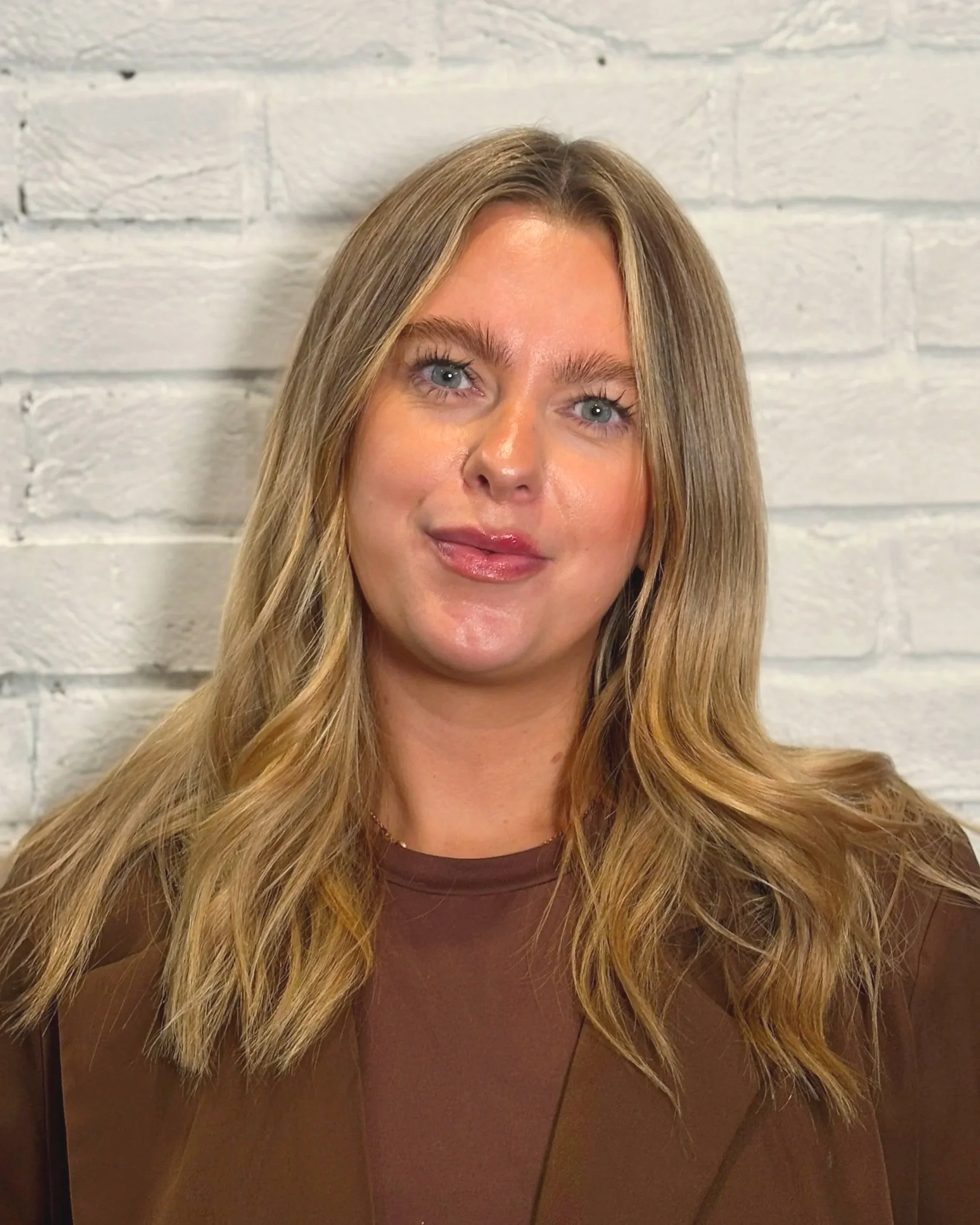 A woman with blonde hair and blue eyes wearing a pinstripe blazer over a grey top standing in front of a mirror in an office setting.