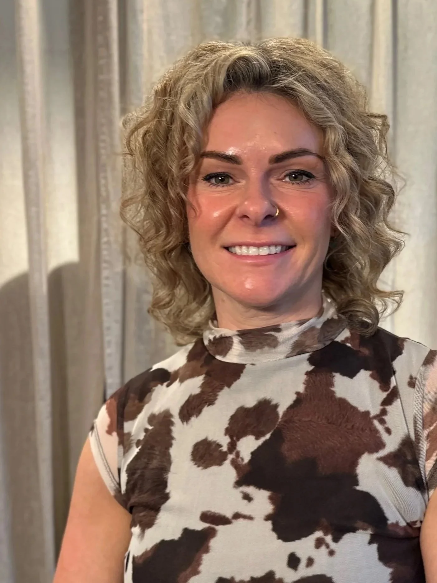 A woman with curly blonde hair smiling, wearing a sleeveless cow-print top and a black belt with a gold GG buckle, standing in front of light-colored curtains.
