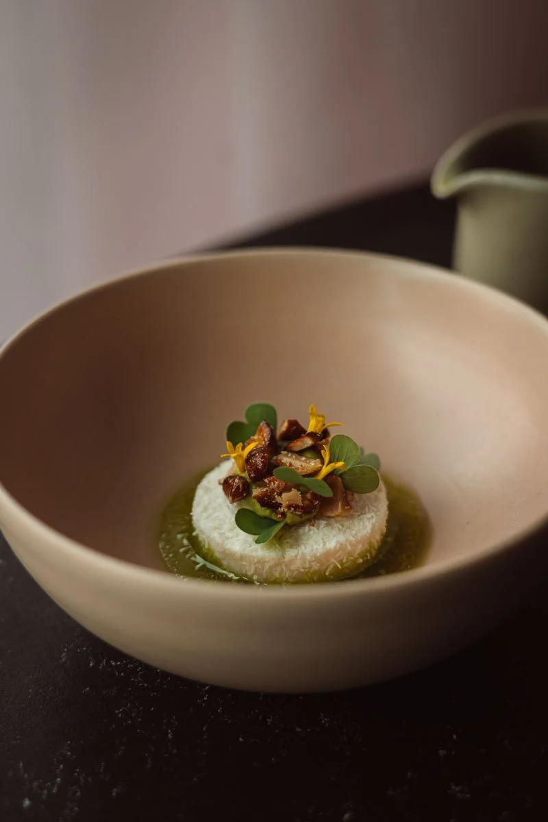 A gourmet dish with a round white base topped with chopped nuts, microgreens, and yellow edible flowers, served in a beige bowl with green sauce. Restaurant Putaine, Rotterdam, The Netherlands