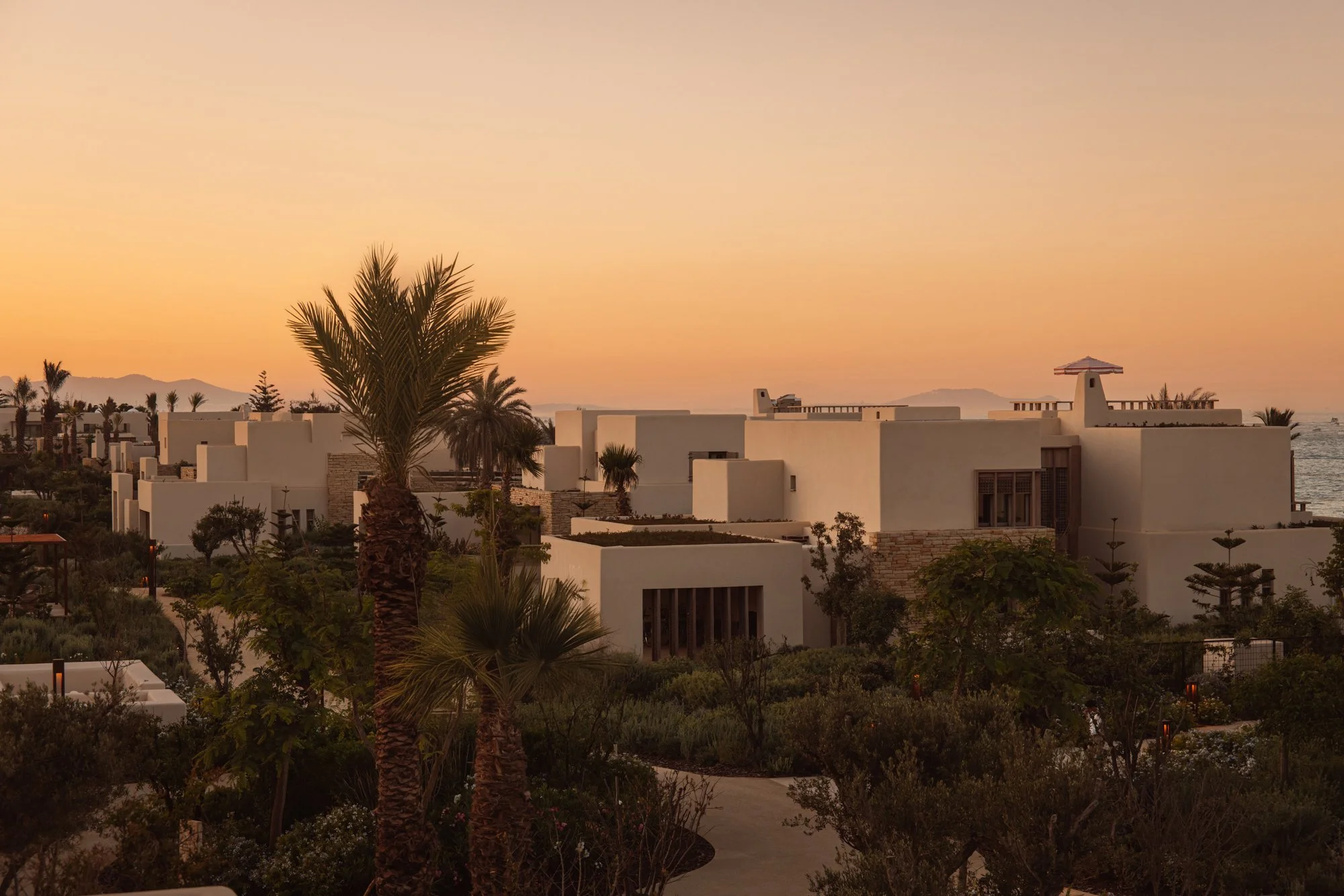 Sunset over modern white buildings with palm trees in a landscaped garden near the coast. Royal Mansour Tamuda Bay Morocco. Photography Sophia van den Hoek