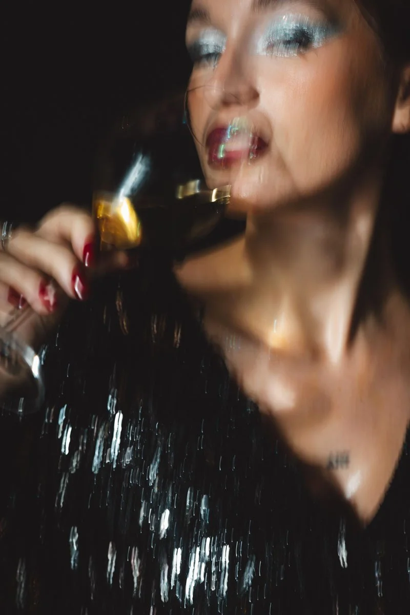 A woman with glamorous makeup, including blue eyeshadow and red lipstick, holds a glass of dark liquid with ice. The image appears to be distorted or blurred. Restaurant Putaine, Rotterdam, The Netherlands