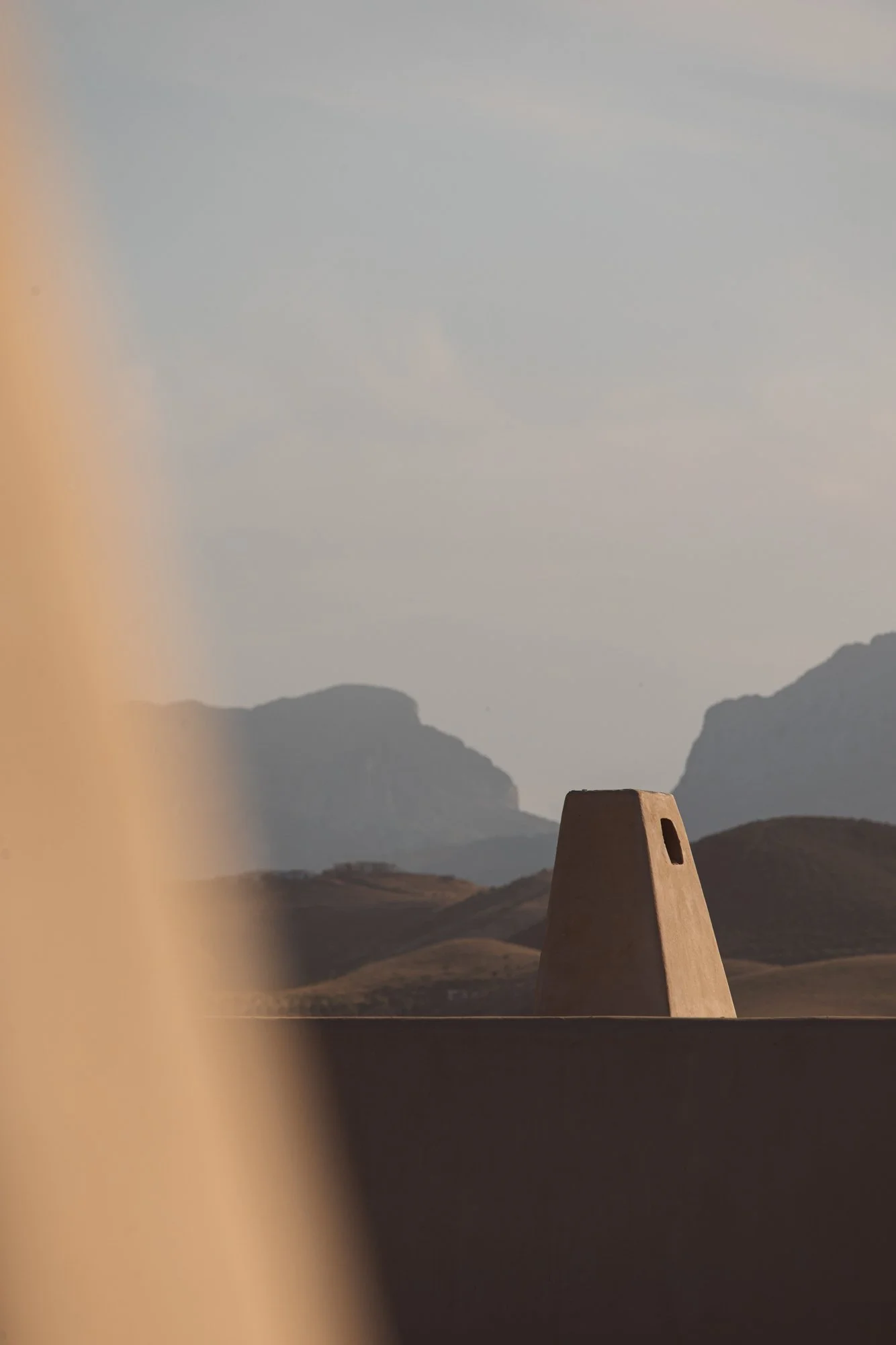 A desert landscape with hills and mountains in the background, a beige structure with a small window in the foreground, and a soft, overcast sky. Royal Mansour Tamuda Bay Morocco. Photography Sophia van den Hoek