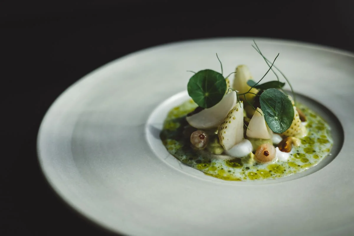 Gourmet dish with white vegetables, green leaves, and sauce on a white plate, dark background. Restaurant Putaine, Rotterdam, The Netherlands