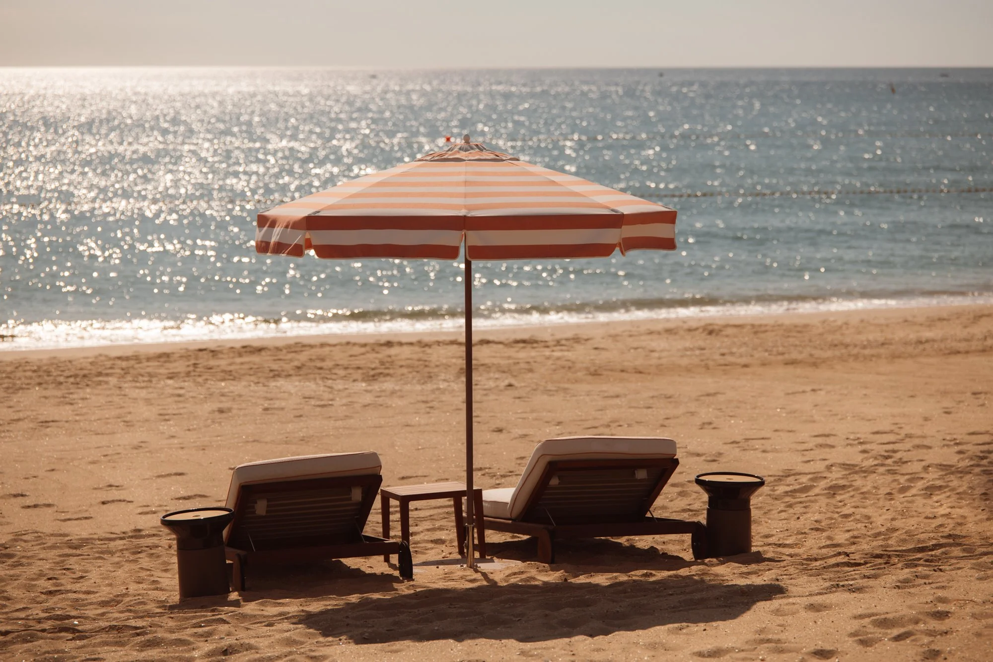 Two empty lounge chairs with cushions under a red and white striped beach umbrella on a sandy beach near the ocean with sparkling water in the background. Royal Mansour Tamuda Bay Morocco. Photography Sophia van den Hoek
