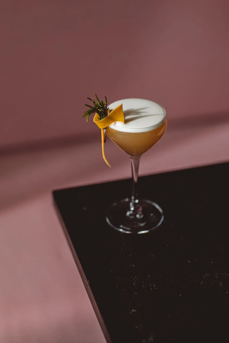 A cocktail drink in a coupe glass garnished with a lemon twist and a sprig of rosemary, placed on a black surface against a pink background. Restaurant Putaine, Rotterdam, The Netherlands