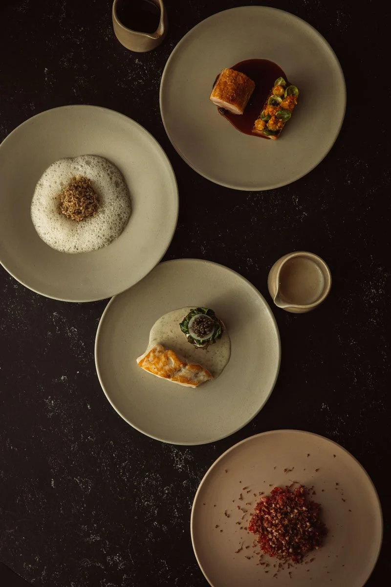 A top-down view of five plates of gourmet food on a dark table, each with a different dish, and a small pitcher of sauce or dressing. Restaurant Putaine, Rotterdam, The Netherlands Photography Sophia van den Hoek