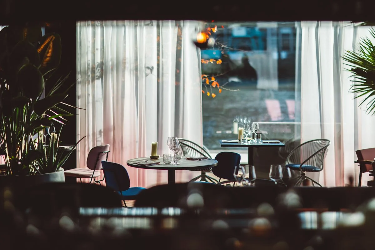 Empty restaurant with tables set with glassware, candles, and utensils, and large windows with curtains, decorated with plants and outdoor scenery. Restaurant Putaine, Rotterdam, The Netherlands