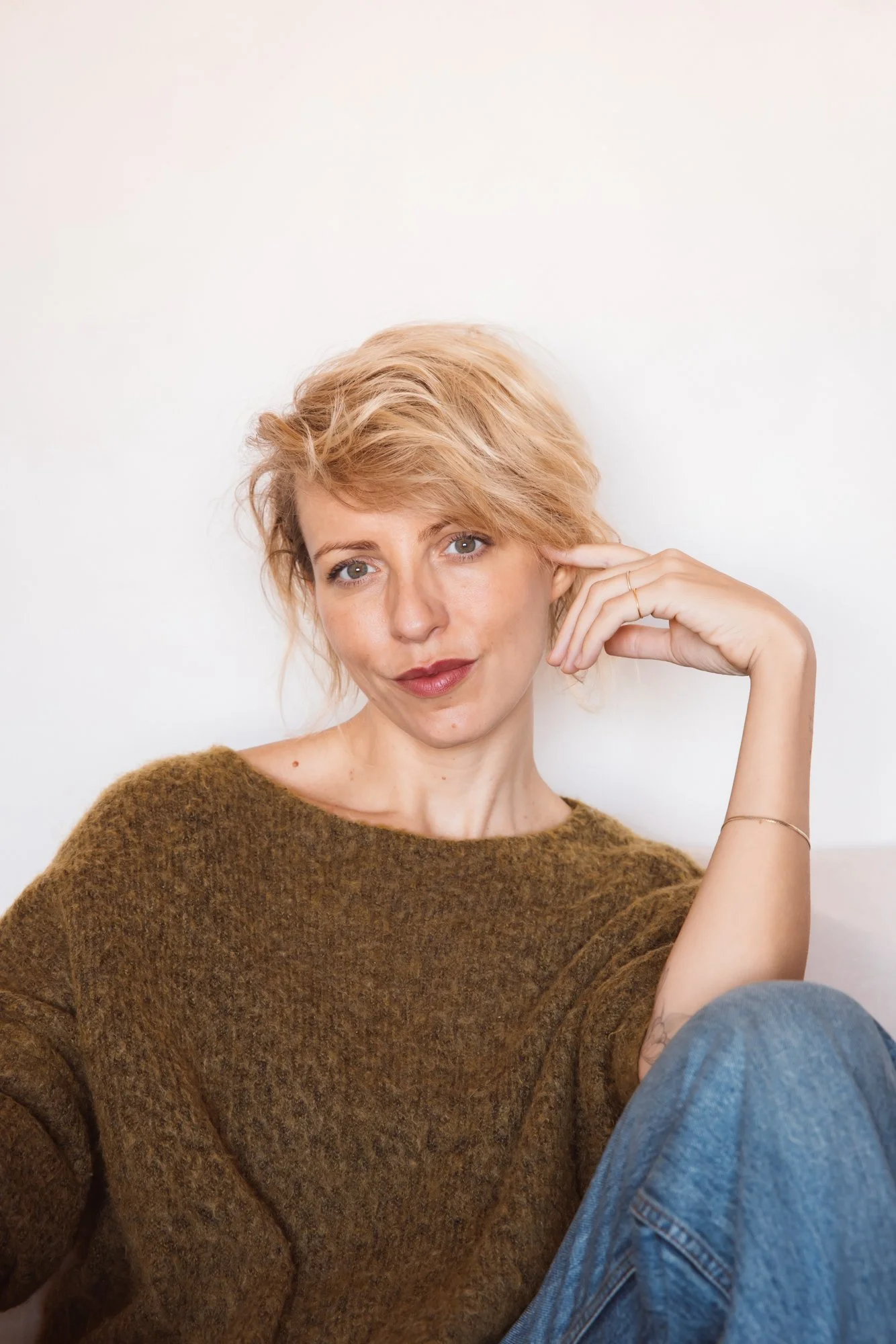 A woman with blond hair and light skin sitting against a plain white wall, wearing a brown sweater and blue jeans, looking at the camera with her hand resting near her face. Sophia van den Hoek, Studio Unfolded