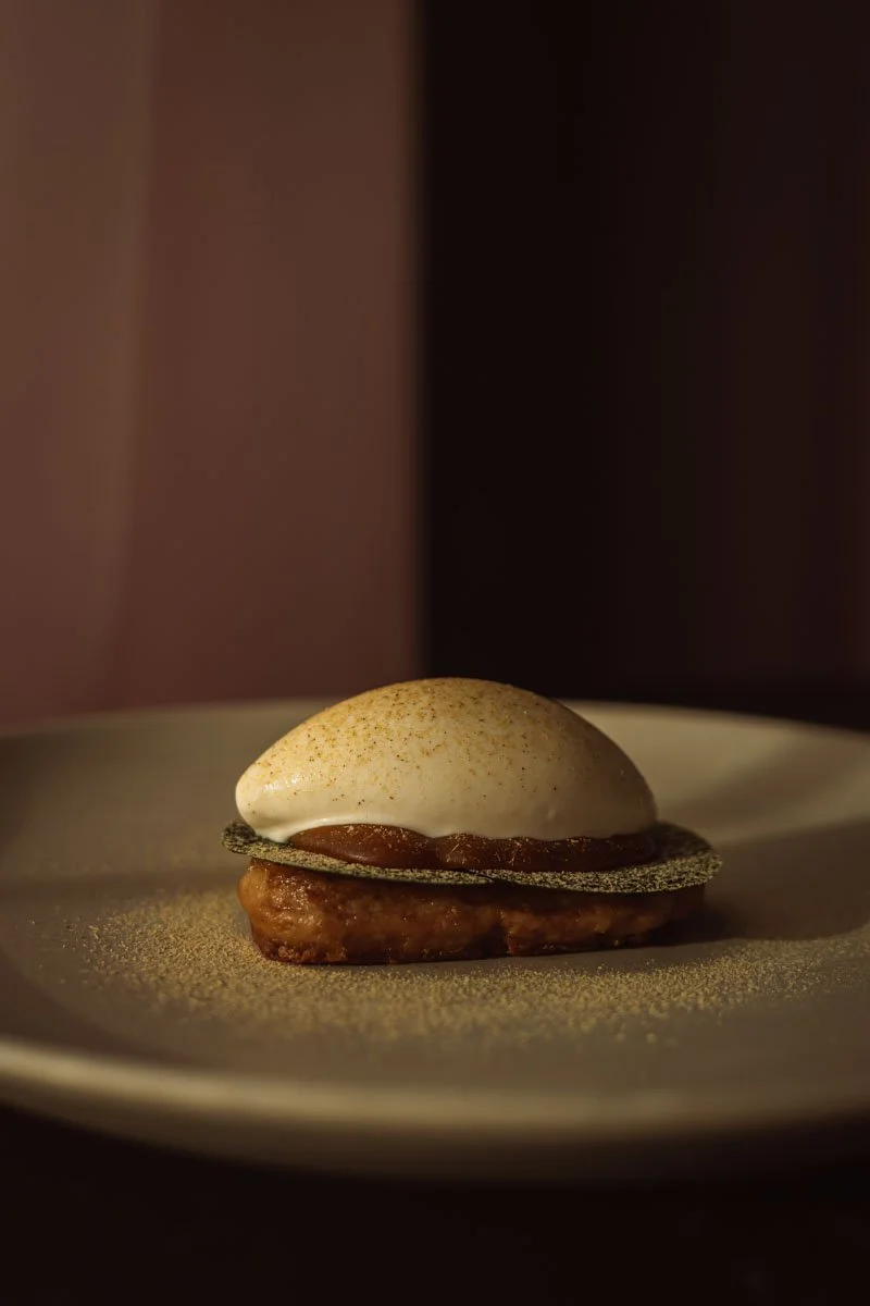 A gourmet dessert featuring a scoop of vanilla ice cream served on a thin, black, crispy wafer, resting on a caramelized piece of fruit, all presented on a simple white plate.Restaurant Putaine, Rotterdam, The Netherlands Photo by Sophia van den Hoek