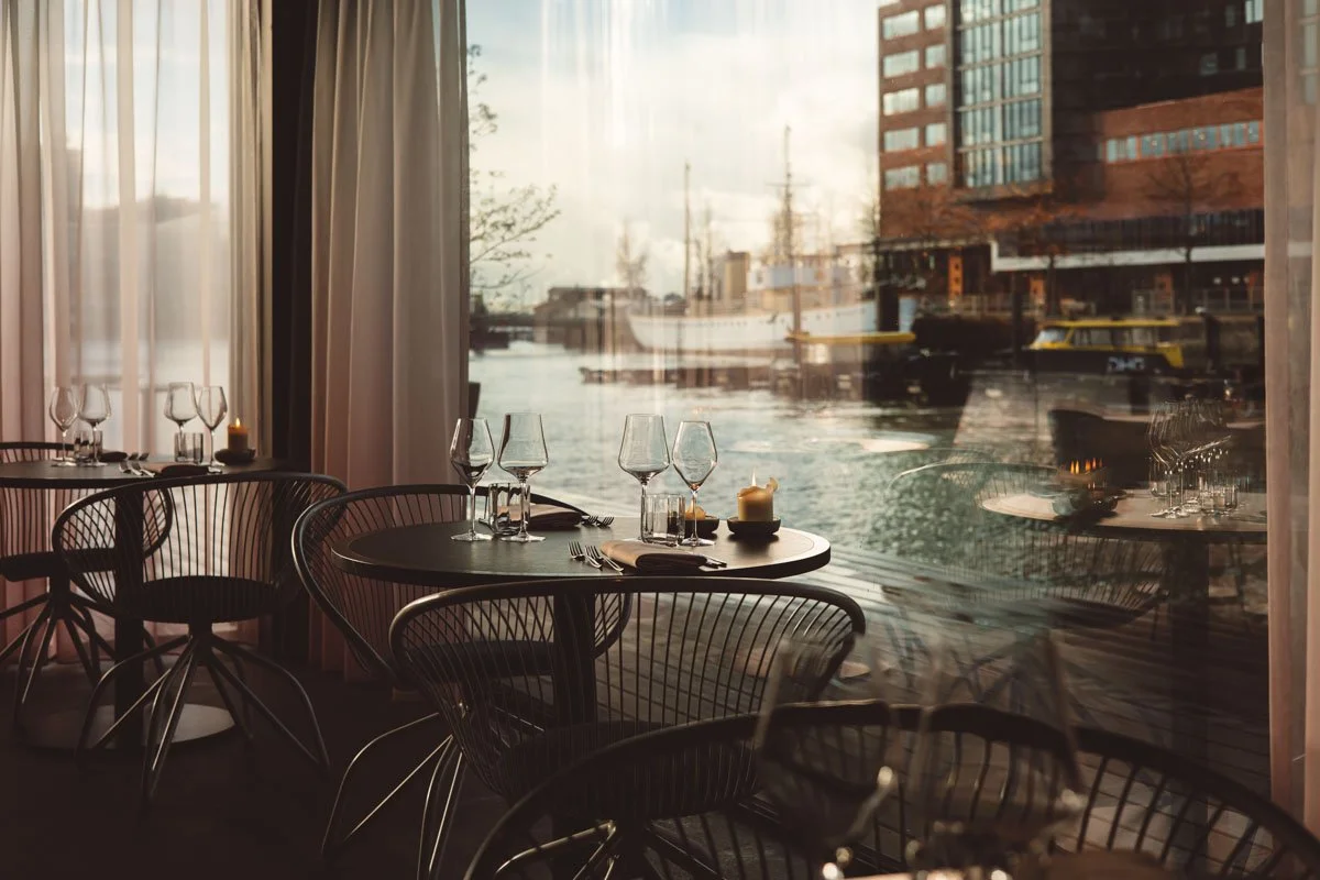 Empty restaurant with black tables and chairs next to large windows overlooking a river and city buildings. Restaurant Putaine, Rotterdam, The Netherlands