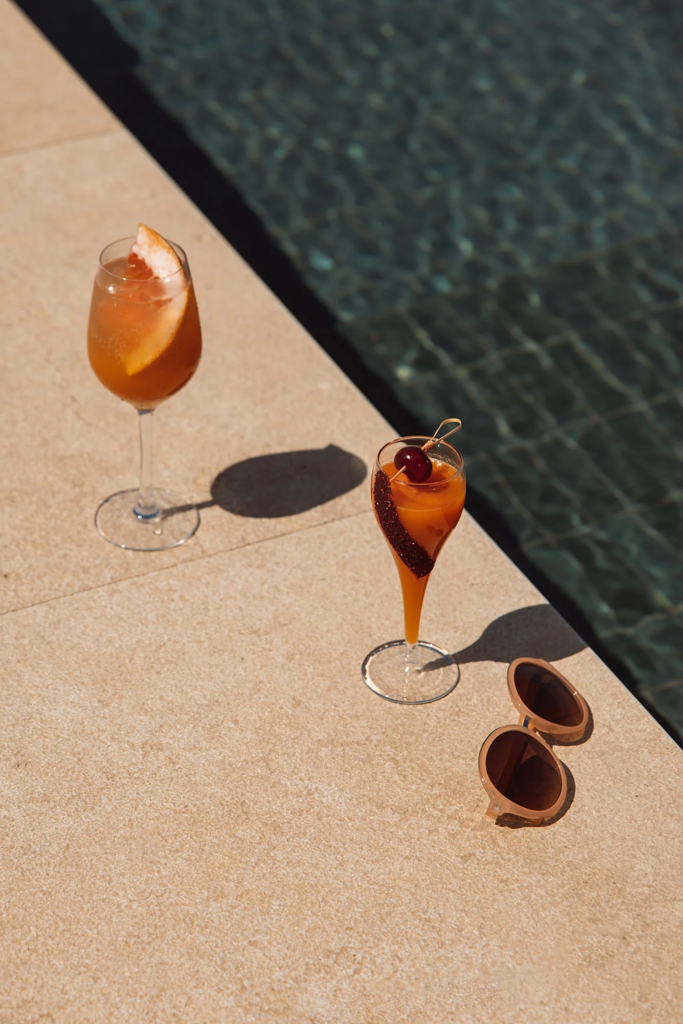 Two cocktails on a poolside surface, with sunglasses nearby and a swimming pool in the background. Royal Mansour Tamuda Bay Morocco. Photography Sophia van den Hoek