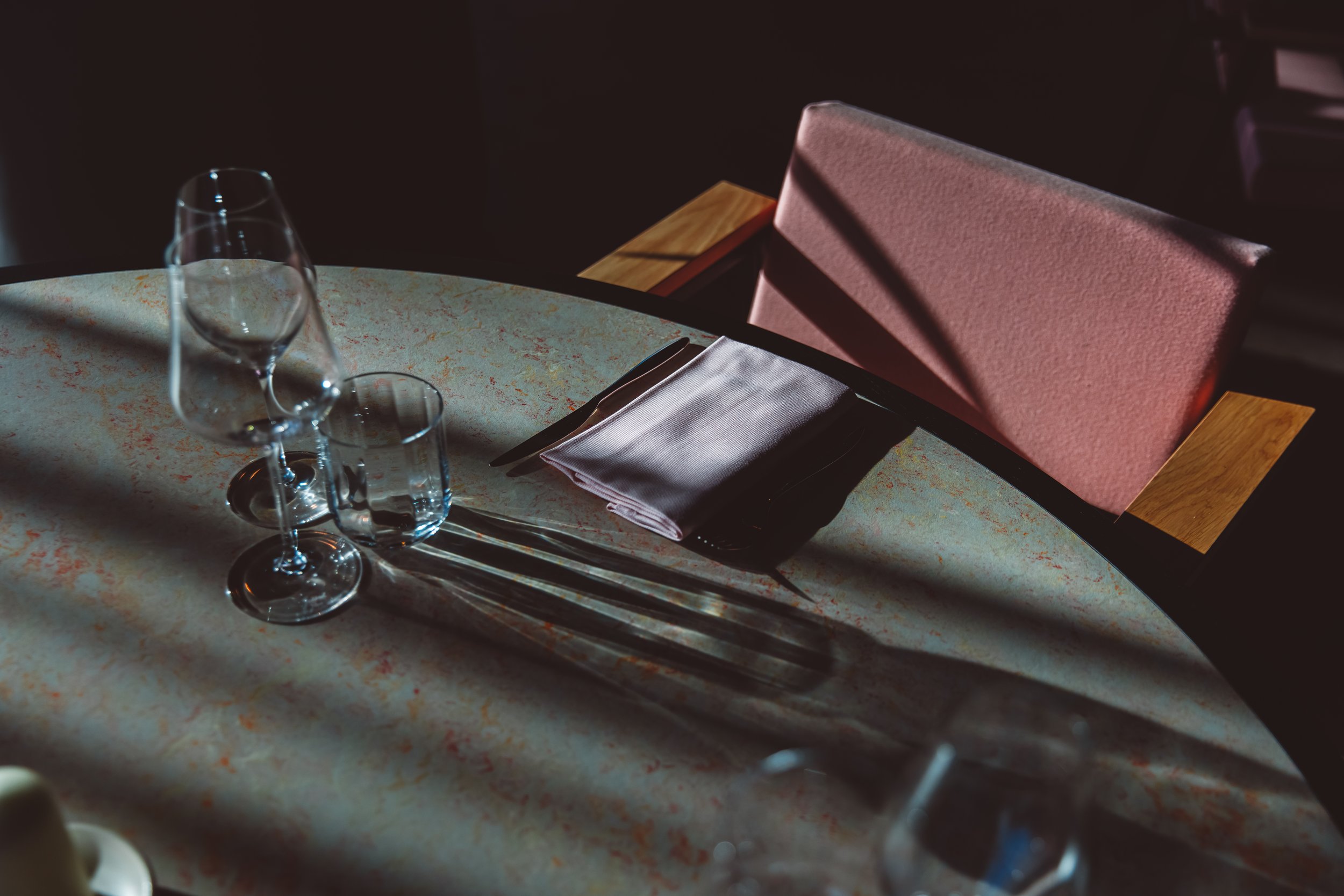 Empty restaurant table set with a wine glass, a water glass, a folded napkin, and utensils, with pink upholstered chairs and warm lighting creating a cozy atmosphere. Restaurant Putaine, Rotterdam, The Netherlands