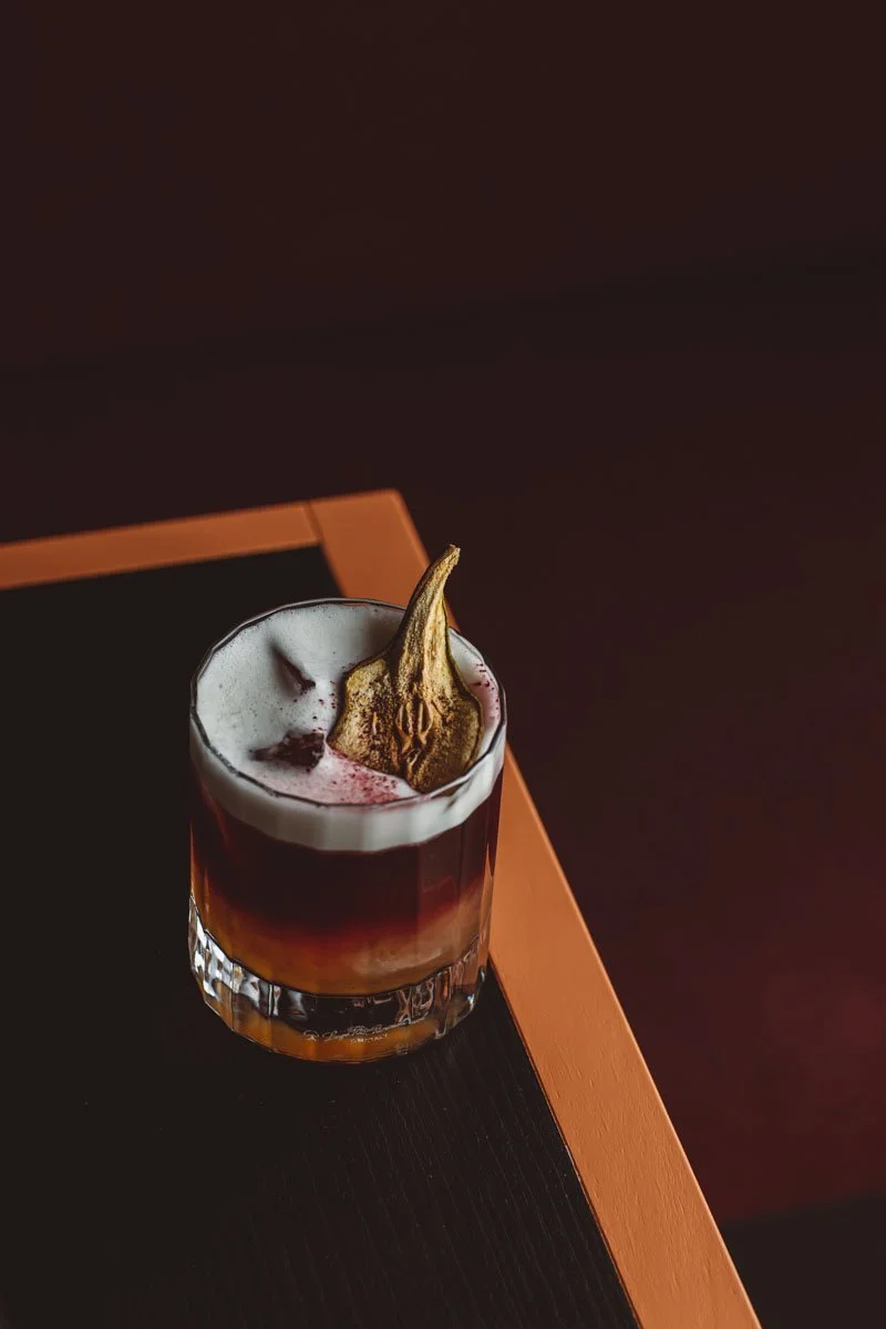 A cocktail in a clear glass with ice, topped with foam and a peacock feather, on a dark wooden table with a wooden edge. Restaurant Putaine, Rotterdam, The Netherlands Photography Sophia van den Hoek