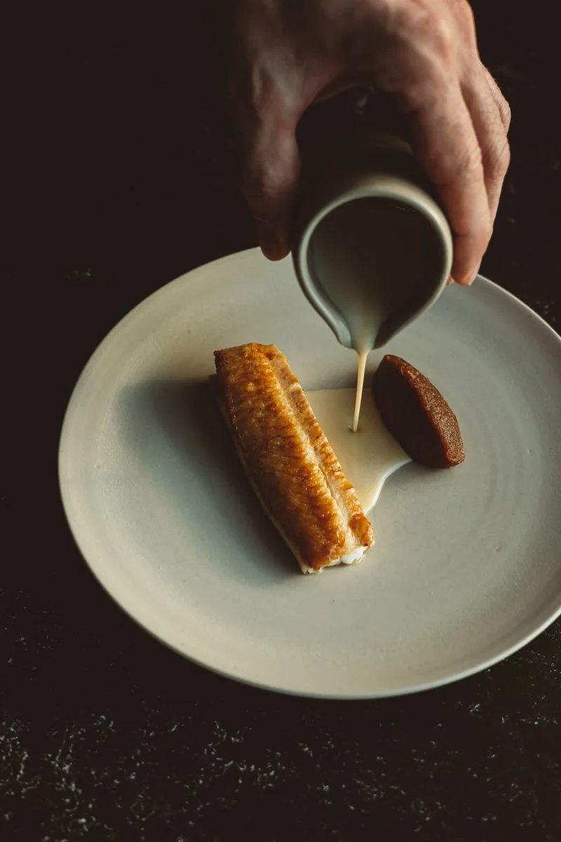 A hand pouring cream or sauce onto a piece of fried fish, with a cookie on a white plate. Restaurant Putaine, Rotterdam, The Netherlands