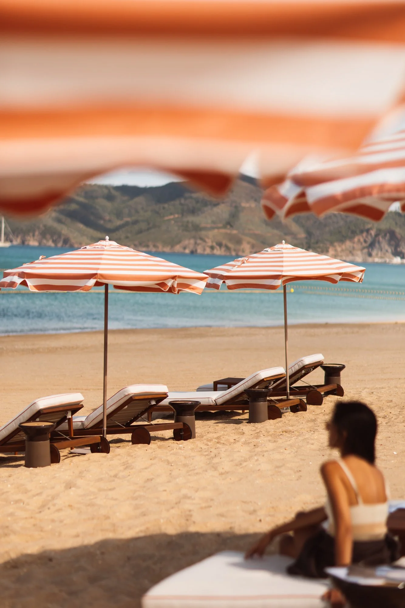 Empty beach with striped orange and white umbrellas, lounge chairs, and a person sitting in the foreground, with a calm body of water and mountains in the background. Royal Mansour Tamuda Bay Morocco. Photography Sophia van den Hoek