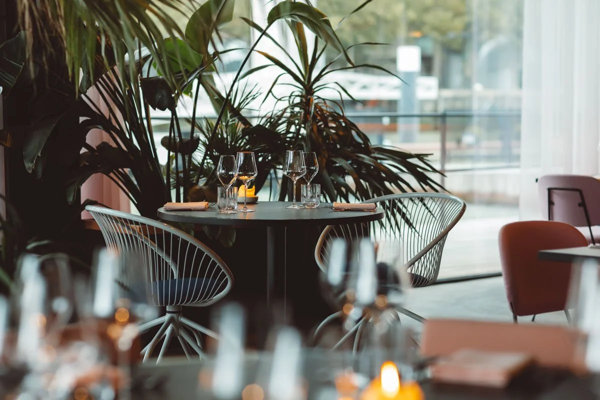 Empty restaurant table set with wine glasses, water glasses, napkins, and silverware, surrounded by modern chairs and large green plants near a window. Restaurant Putaine, Rotterdam, The Netherlands