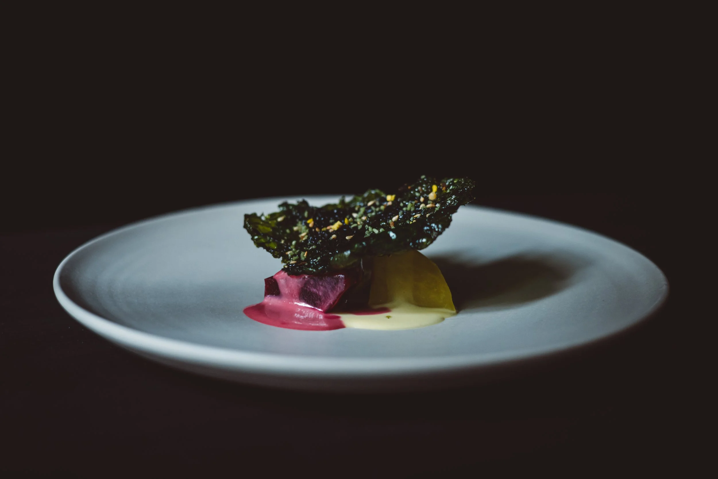 A gourmet dish featuring a piece of pink and yellow meat or vegetable, topped with a large crispy green seaweed chip, served on a white plate with pink and yellow sauces, against a dark background. Restaurant Putaine, Rotterdam, The Netherlands
