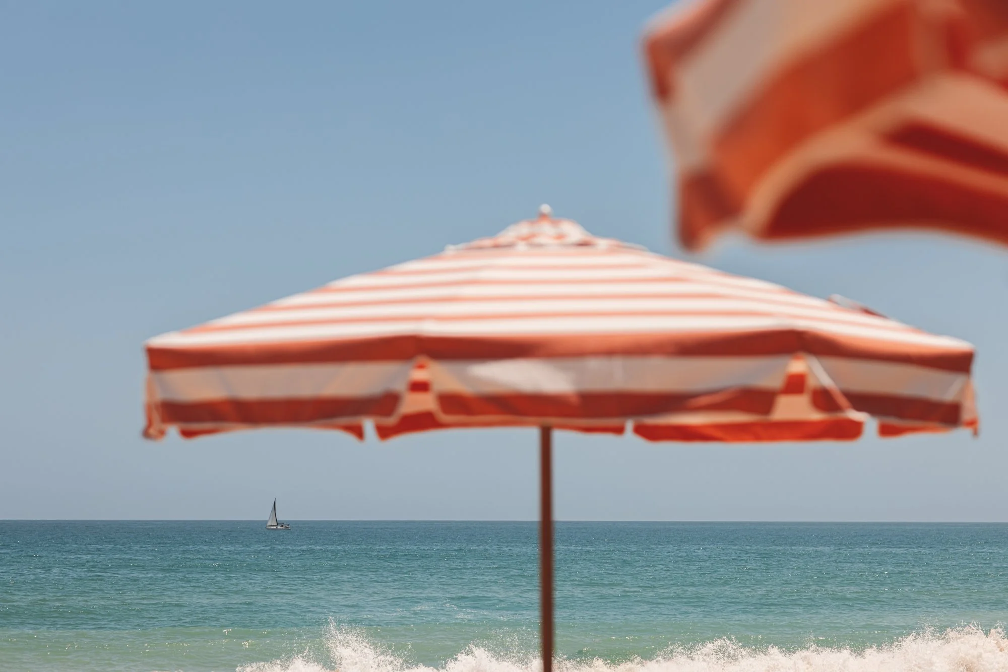 Beach scene with a large striped orange and white umbrella in the foreground and a sailboat on the ocean in the background. Royal Mansour Tamuda Bay Morocco. Photography Sophia van den Hoek