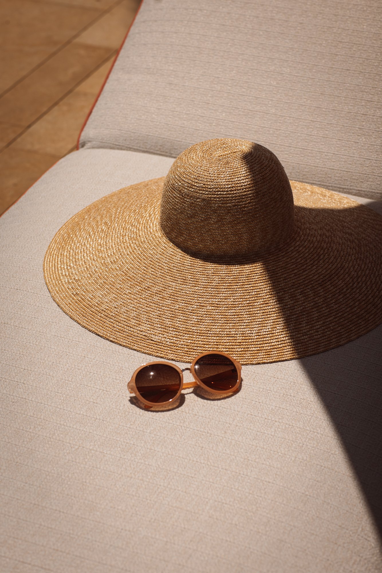 A large straw sun hat and a pair of round sunglasses rest on a beige cushioned outdoor chair, with sunlight casting shadows. Royal Mansour Tamuda Bay Morocco. Photography Sophia van den Hoek