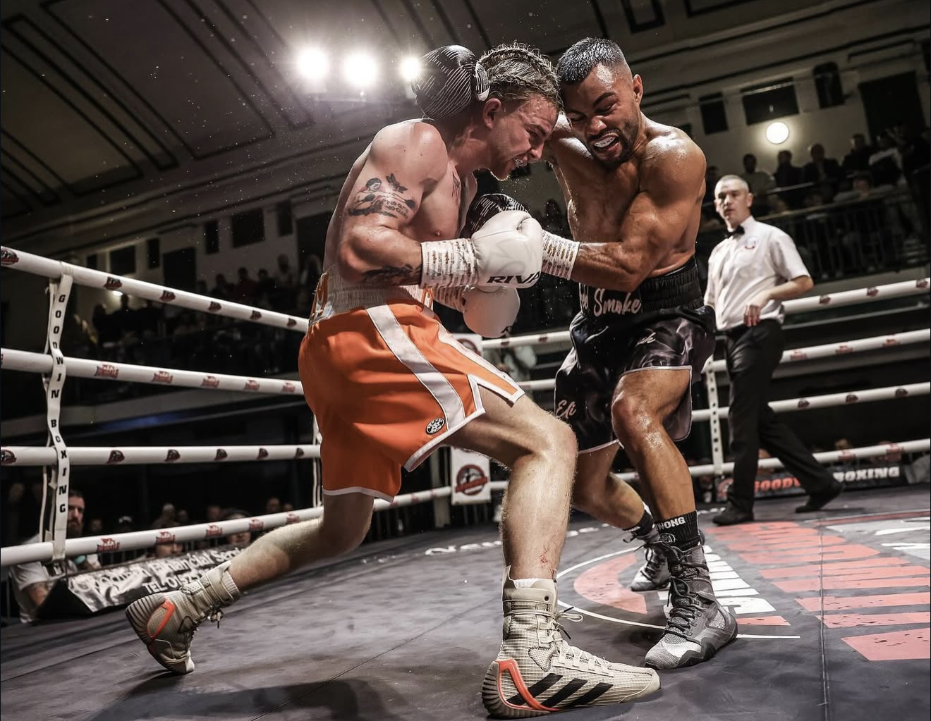 Two male boxers fighting intensely in a boxing ring, with a referee watching in the background. One boxer is wearing orange shorts and the other black, both wearing boxing gloves. The boxer in orange appears to be throwing a punch while the other is defending.