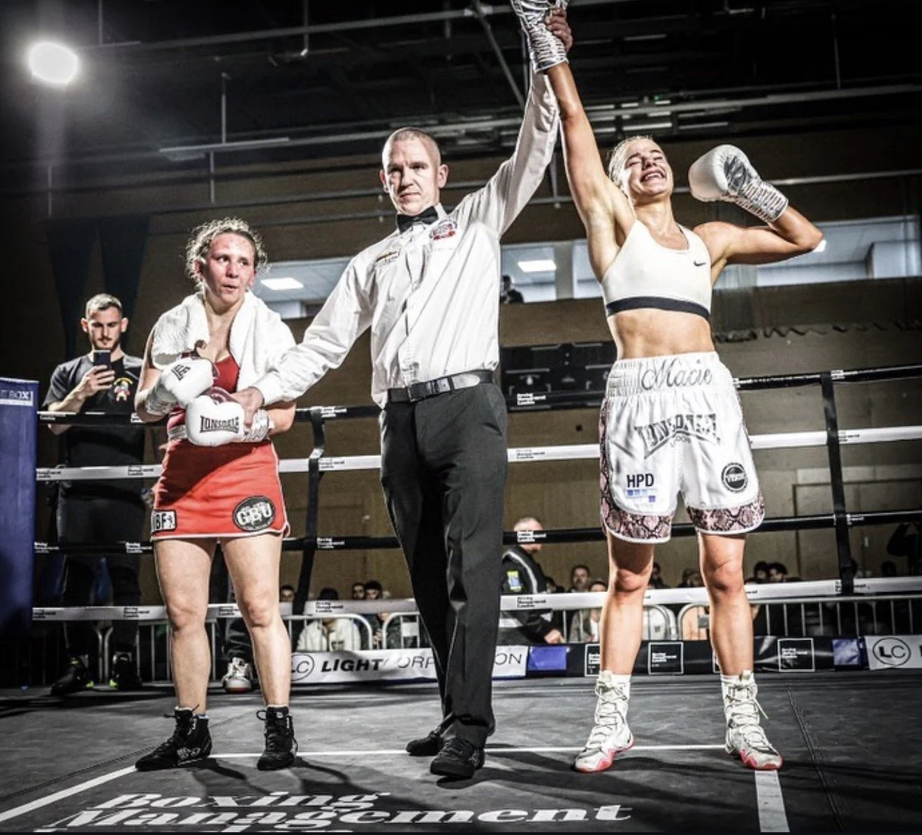 Boxer with blonde hair celebrating victory with raised arm, wearing white shorts and sports bra, in boxing ring. Opponent woman in red shorts appears disappointed. Referee in black and white shirt is holding their hands, in sports hall with spectators.