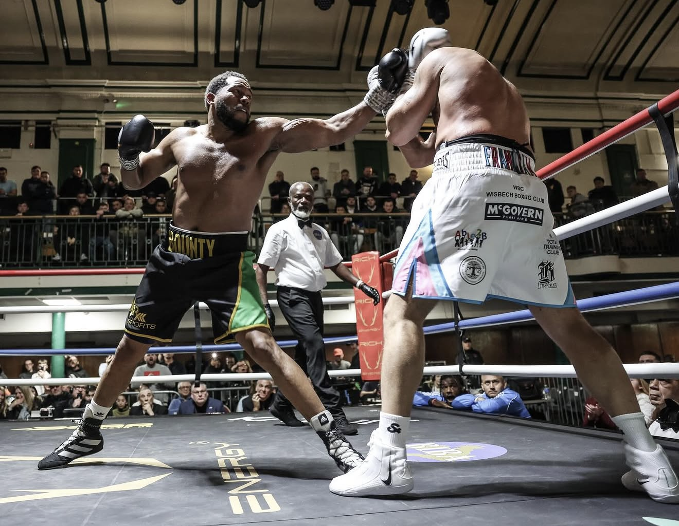 Two boxers in a boxing ring engaged in a fight, with one delivering a punch to the other's face. Spectators and a referee are visible in the background.