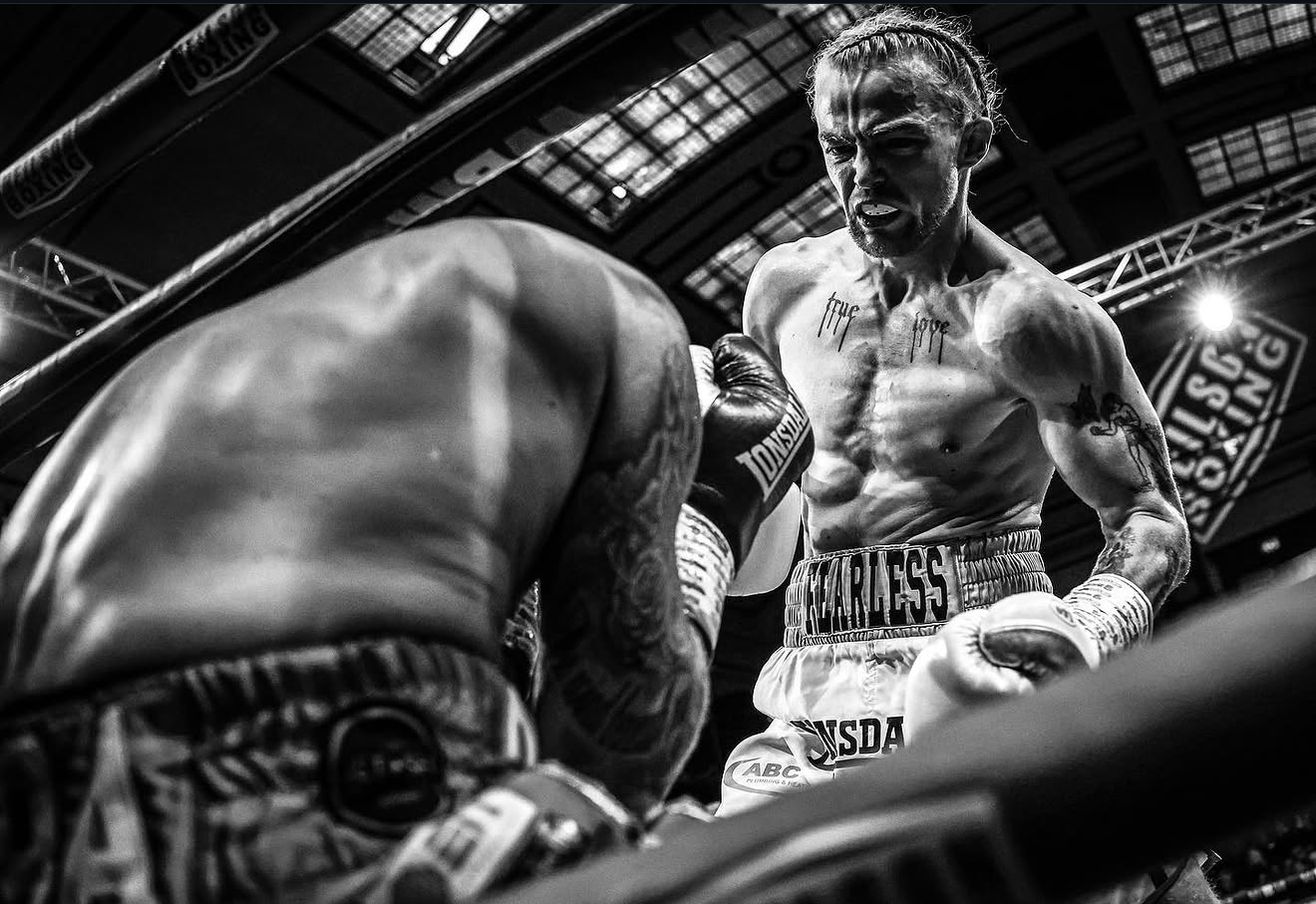 A black and white photograph of two male boxers in a boxing ring, one with a beard, standing and staring intensely at his opponent. The boxer on the right has visible tattoos on his chest and arms, and is wearing boxing shorts with "FEARLESS" written on the waistband. The other boxer is facing away from the camera, his back and shoulders visible, and wearing boxing gloves and shorts. The background shows the ceiling of the arena with lights and banners.