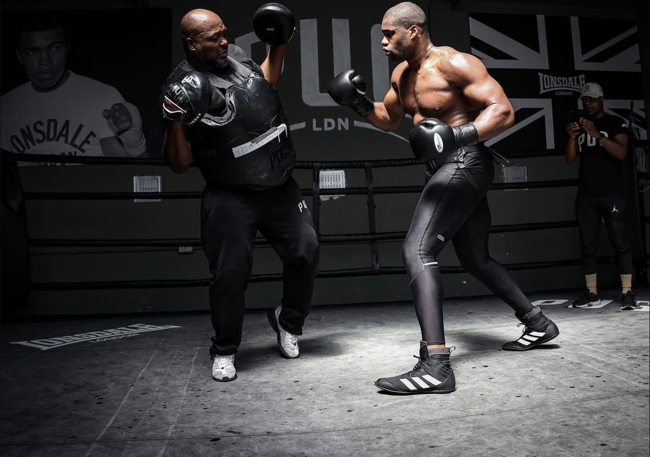 Two boxers training in a boxing ring, with one in black gloves and a shirt holding a focusing shield, and the other shirtless, wearing black tights and boxing shoes, practicing punches. A person in the background is recording with their phone.