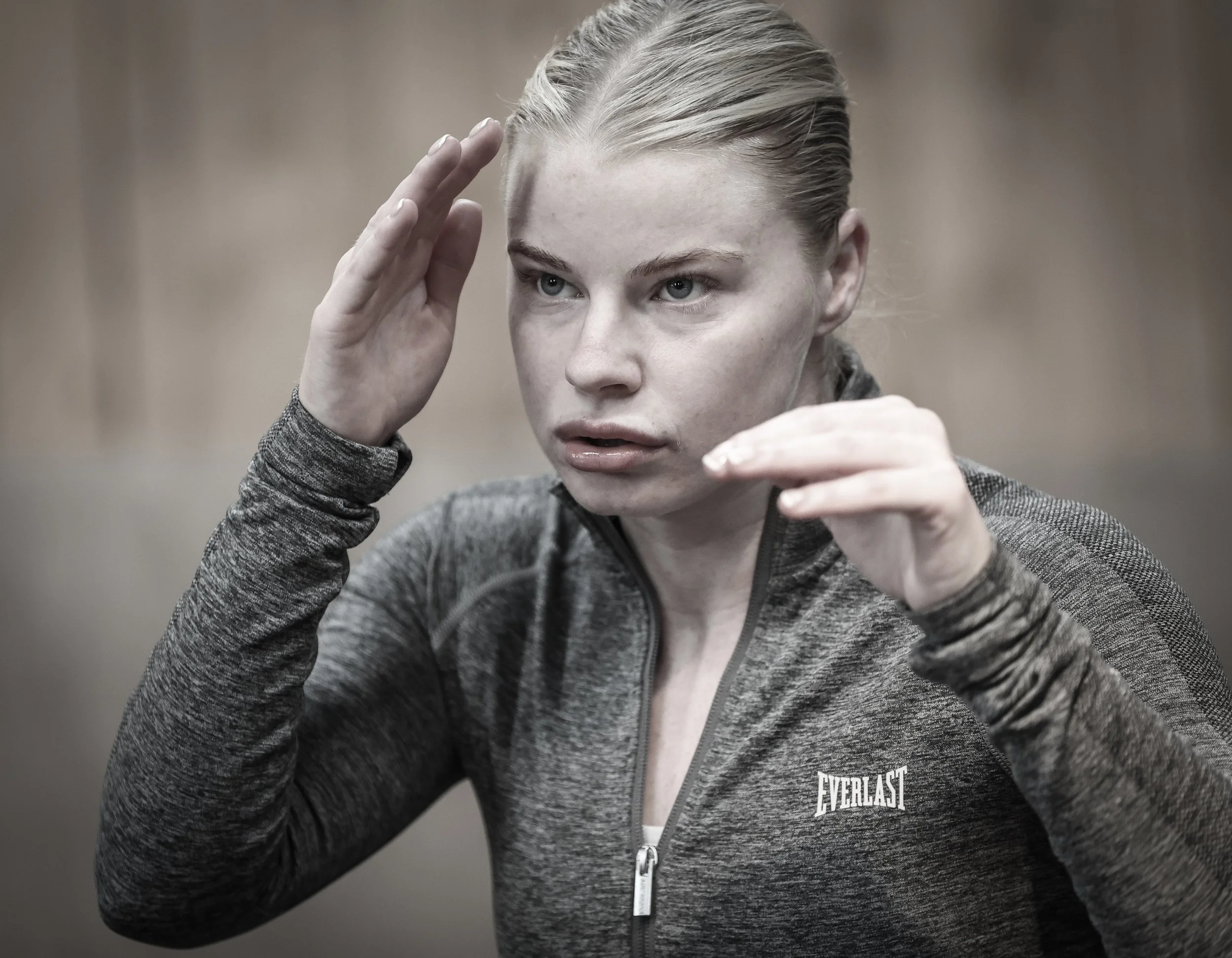 A woman with blonde hair pulled back, wearing a gray athletic zip-up jacket with Everlast logo, practicing boxing, with her hands raised in a defensive stance.