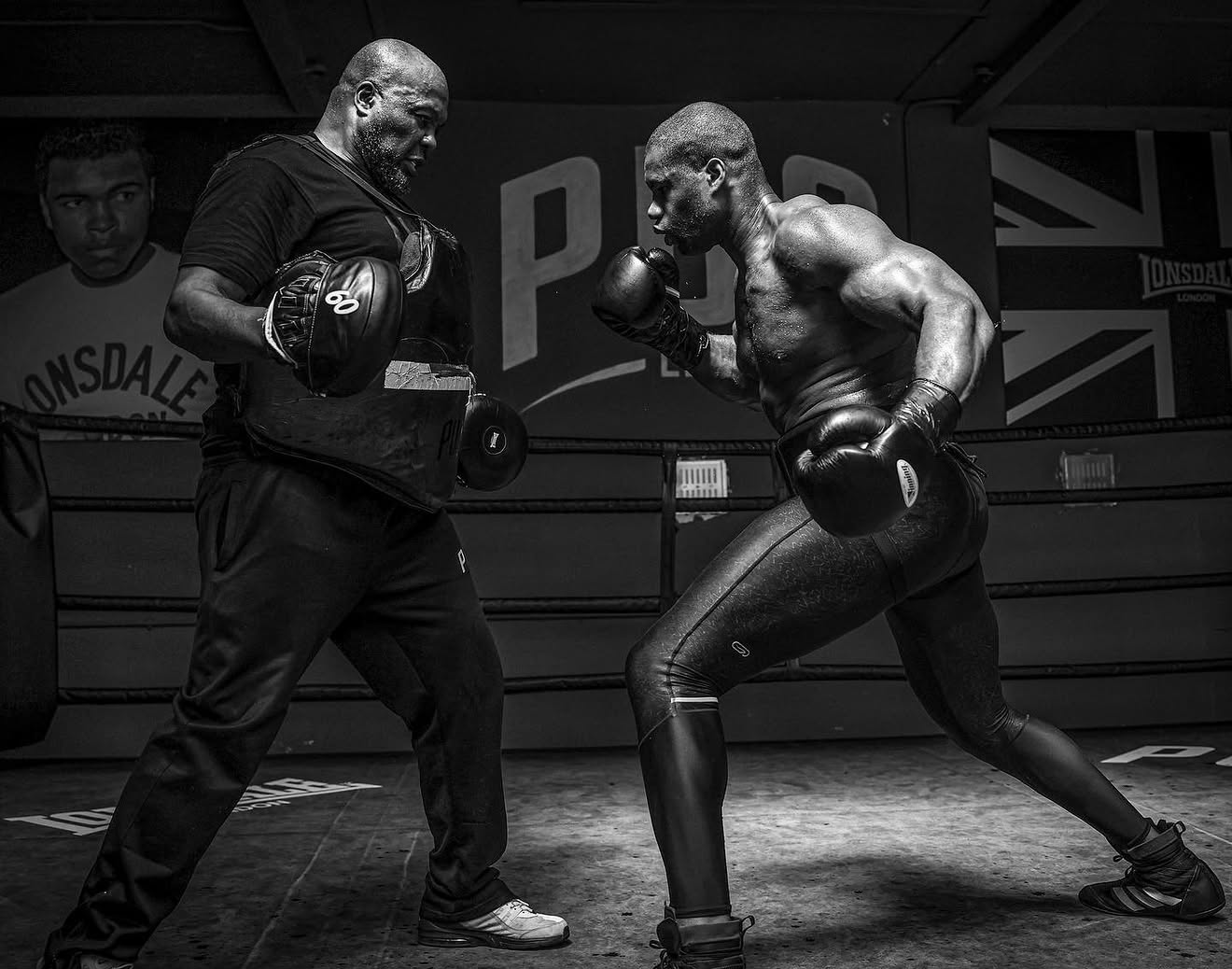 Two male boxers in a boxing ring, sparring with focus and intensity. One is wearing gloves and a black shirt, the other is shirtless with traditional boxing gloves. A poster of a young boxer and the Union Jack flag can be seen in the background.