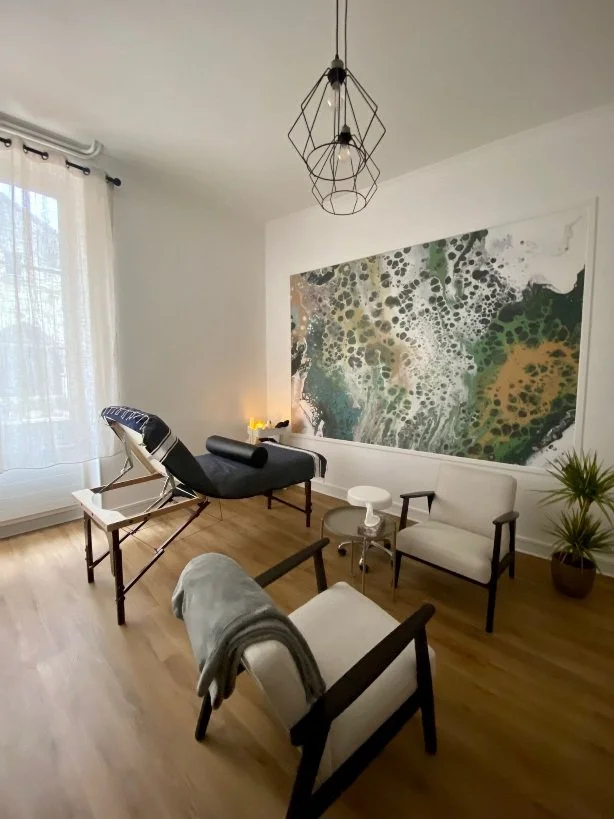 Salon moderne avec canapé beige, fauteuils en bois, tableau abstrait sur le mur, lampadaire vintage, plante en pot, et lumière naturelle venant d'une fenêtre avec rideaux blancs.