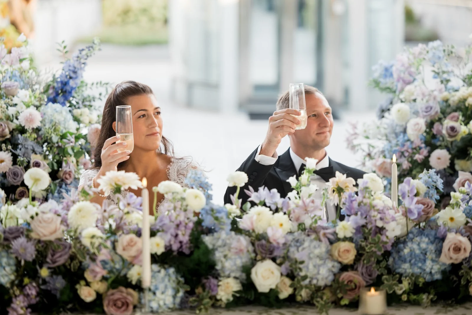 Bride and groom at their wedding reception, sitting behind a floral arrangement, raising glasses of champagne for a toast.