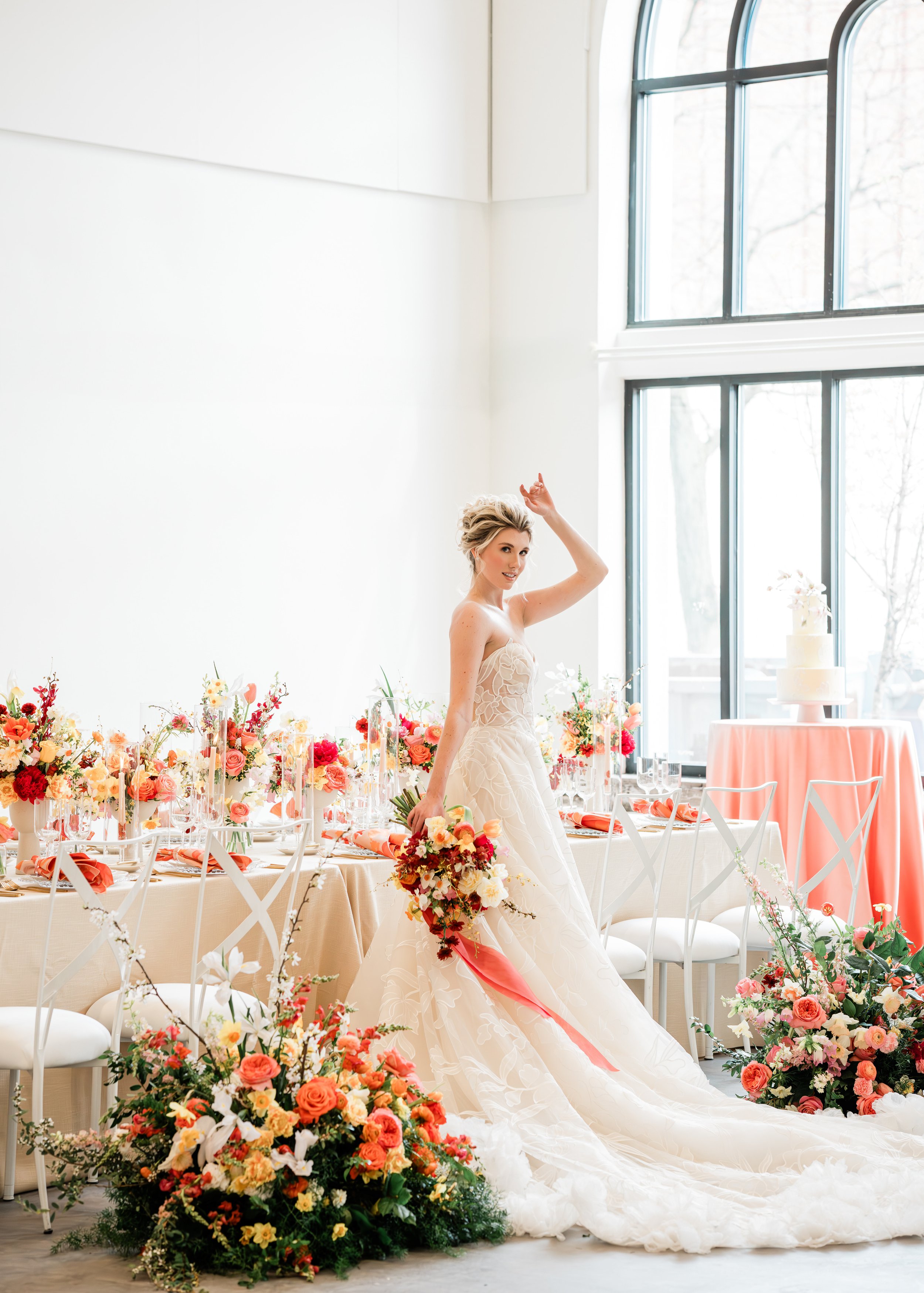A bride in a wedding dress standing in a bright room decorated with colorful flower arrangements, holding a bouquet of flowers, near a table with floral decor and a wedding cake in the background.