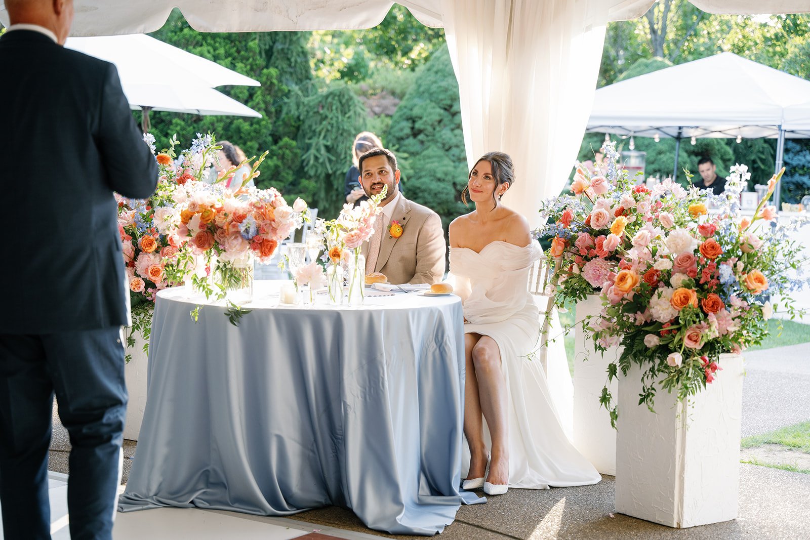 Wedding ceremony with bride and groom seated at a table decorated with colorful flowers, outdoors under a white canopy, with a officiant standing in front, in a garden setting.