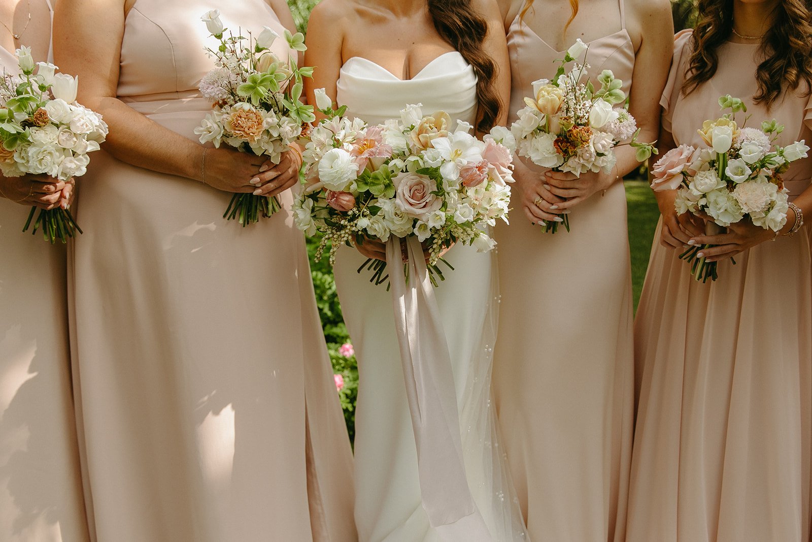 Bridal party standing outdoors, holding bouquets of pink and white flowers with greenery at a wedding.