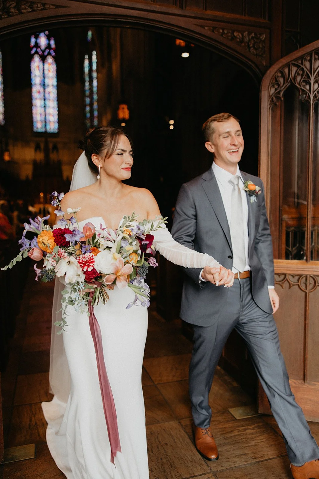A bride and groom holding hands as they walk into a church for their wedding ceremony. The bride is wearing a white off-shoulder gown and holding a large, colorful bouquet of flowers. The groom is dressed in a gray suit with a white shirt and tie, an