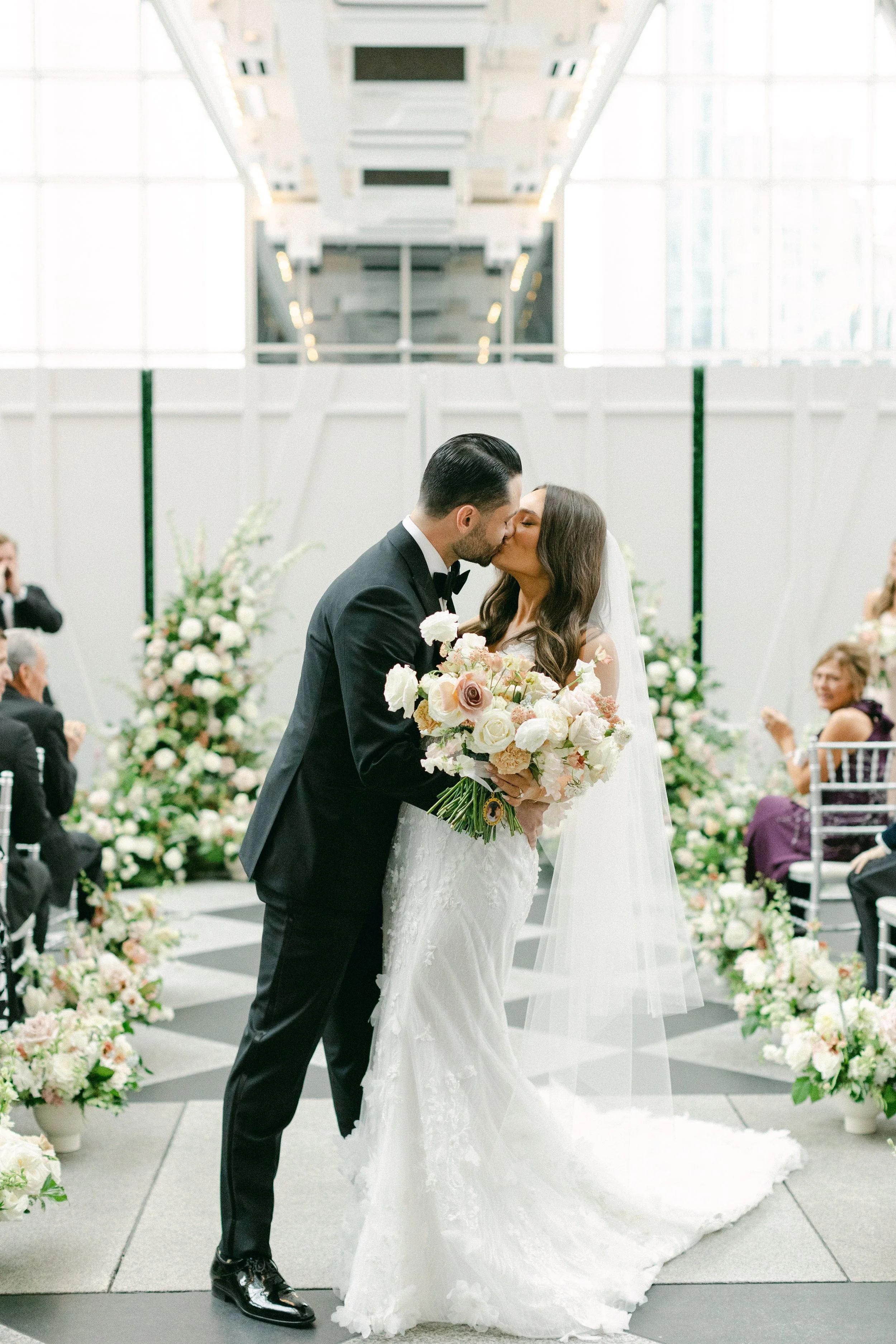Wedding couple at PPG Wintergarden in downtown Pittsburgh kissing at ceremony with large floral backdrop and stunning aisle runner of flowers.