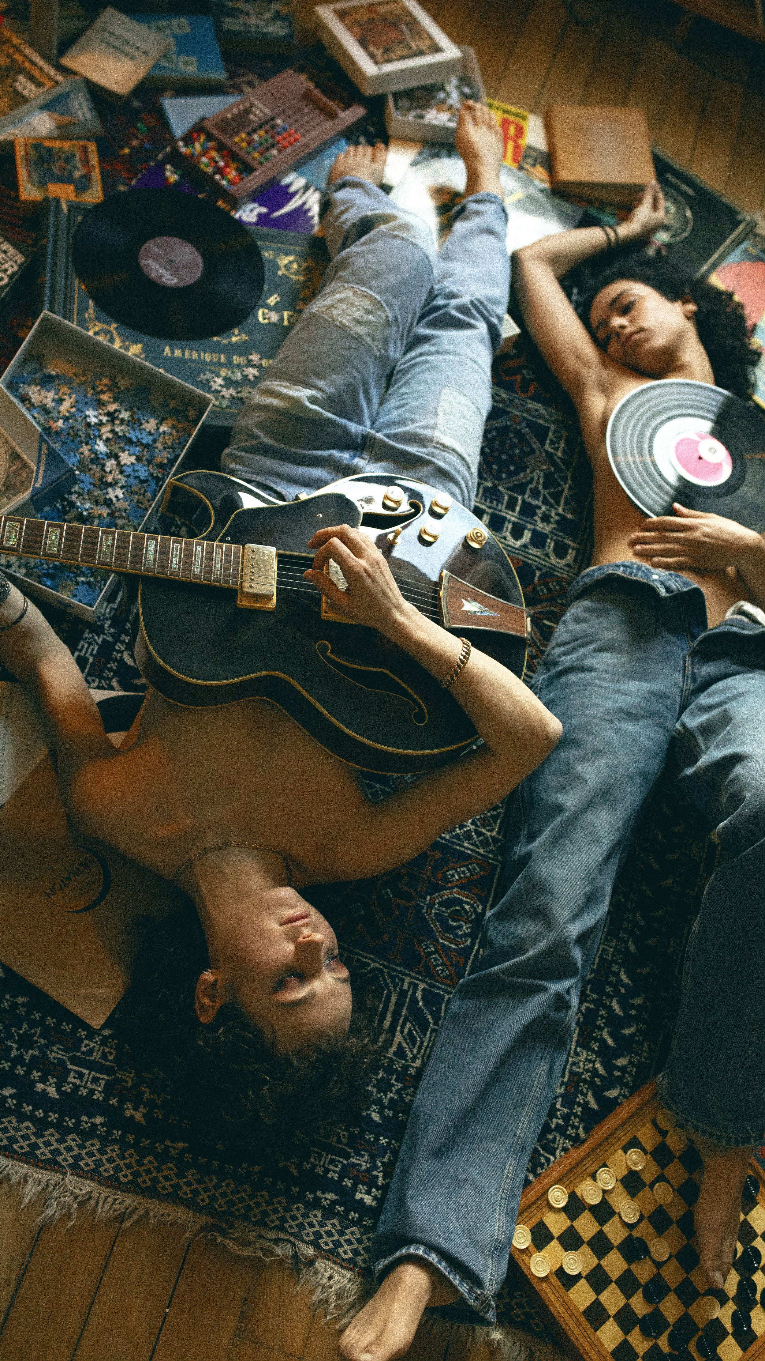 Deux jeunes adultes, un homme et une femme, allongés sur un tapis, entourés de jeux de société, disques vinyles, et une guitare. La femme joue de la guitare, l'homme tient un disque vinyle, tous deux semblent se détendre dans une ambiance conviviale.