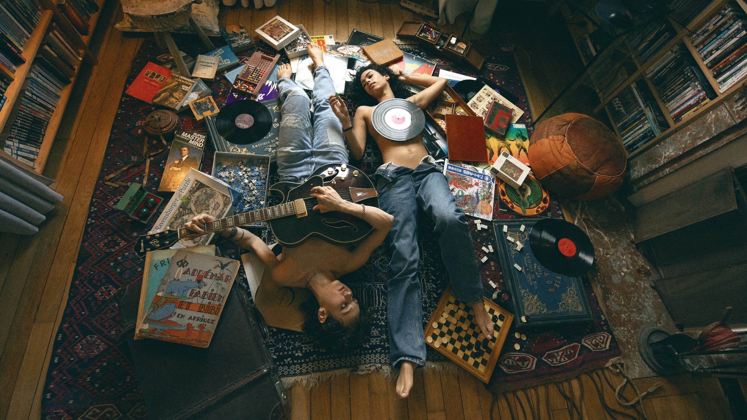 Deux personnes allongées sur un tapis, entourées de livres, vinyles, jeux de société, et autres objets de clutter, dans une ambiance de chambre ou salon.
