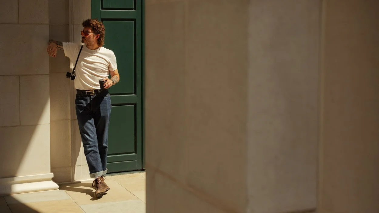 man standing in corridor wearing black leather belt with brass buckle and blue jeans and white tshirt with coffee cup