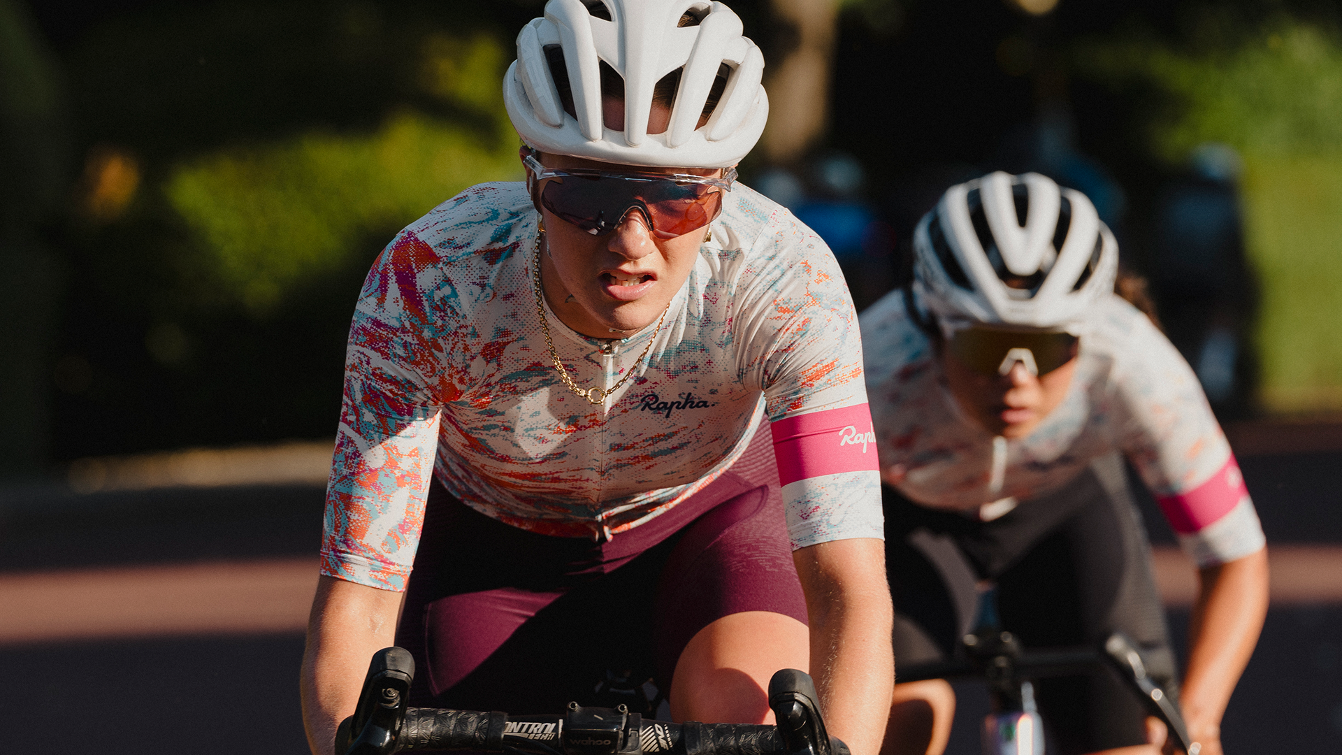 Two female cyclists, wearing white helmets, sunglasses, and colorful cycling jerseys, are riding on a road during daytime with sunlight filtering through trees in the background.