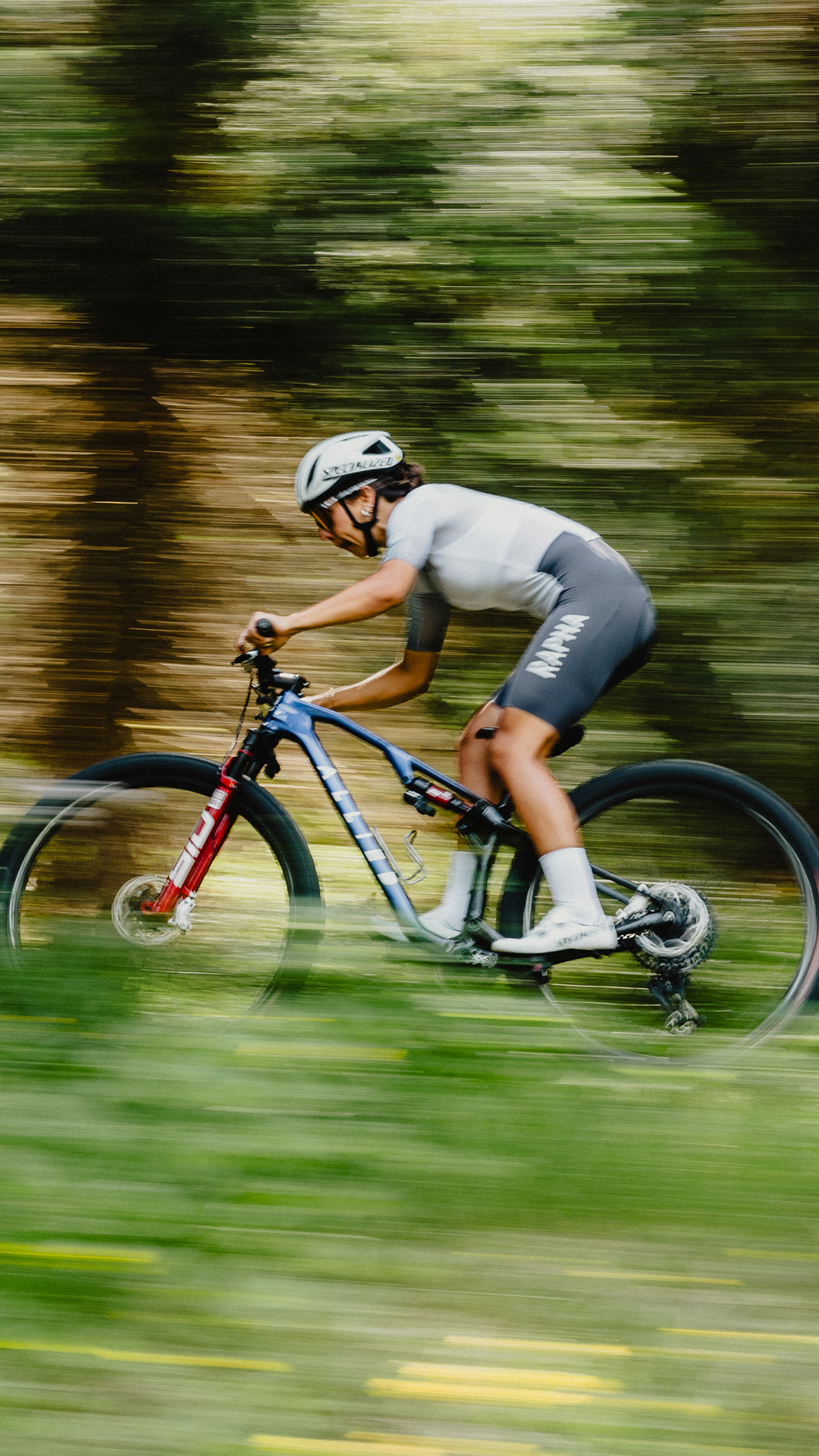 A person riding a mountain bike through a forest, wearing a white helmet and cycling gear, with motion blur indicating fast movement.