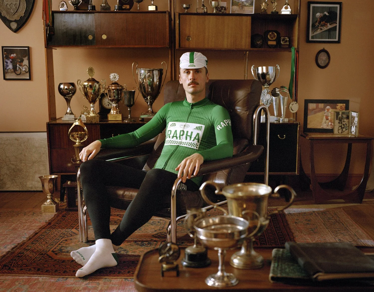 A man dressed in a green Rapha cycling outfit with a matching cap, sitting in a vintage armchair in a room with numerous trophies on shelves behind him, surrounded by framed pictures and medals.