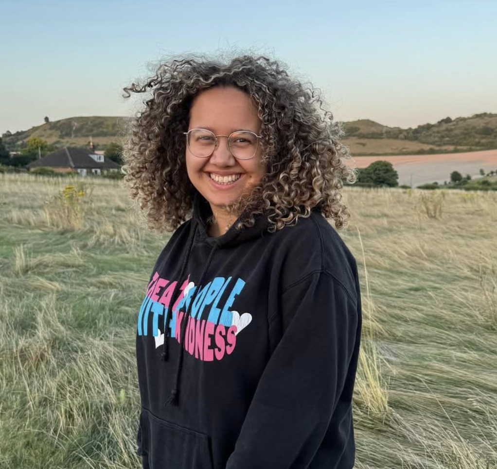 A young woman with curly hair and glasses smiling outdoors in a field with hills and houses in the background.