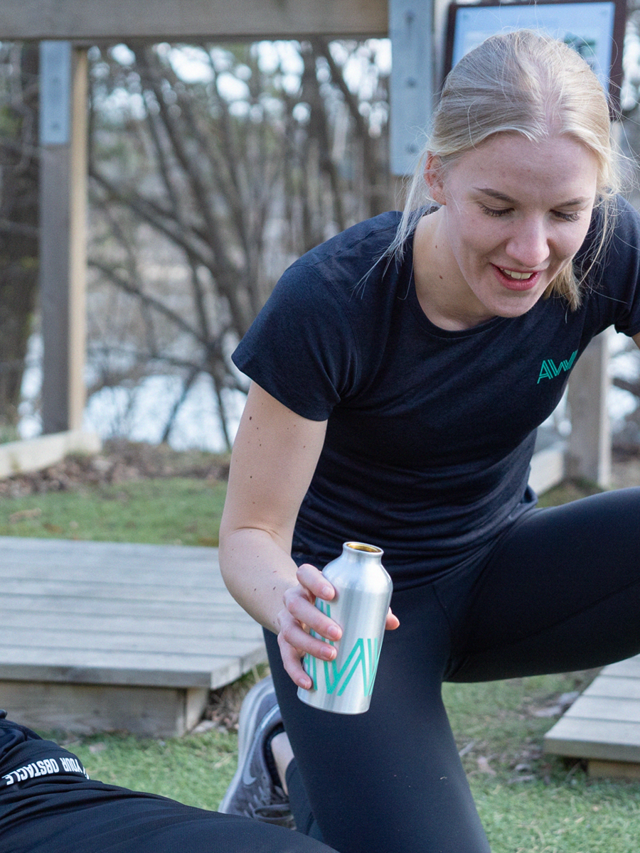 A young woman with blonde hair in a ponytail wearing a black shirt and black pants, holding an opened silver can with a teal design, outdoors with a wooden deck and trees in the background.
