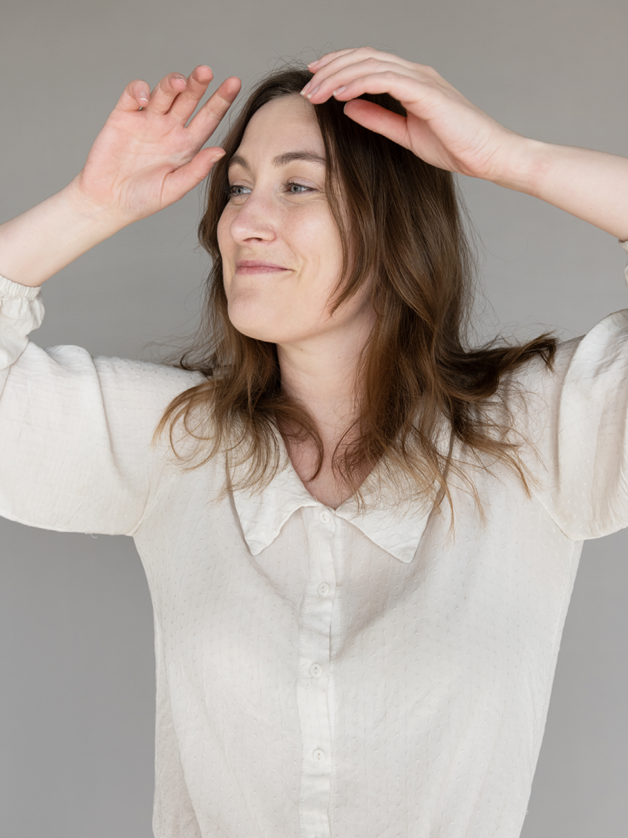 A woman with brown hair wearing a cream-colored blouse, smiling with her eyes closed, hands raised near her head, against a gray background.